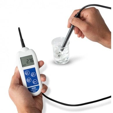 Une personne tenant un Conductimètre 8500 de Thermomètre.fr et un verre d'eau pour mesurer sa conductivité en mS/cm.