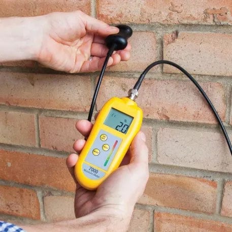 une personne tenant un Testeur d'humidité 7000 avec sonde de Thermometre.fr devant un mur de briques.