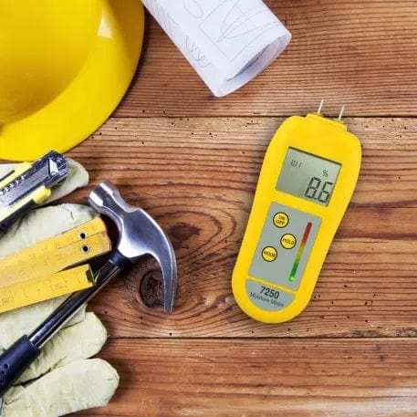 Un Humidimètre jaune pour le bois et les matériaux de construction avec un casque et des outils sur une table en bois. (Marque : Thermometre.fr)